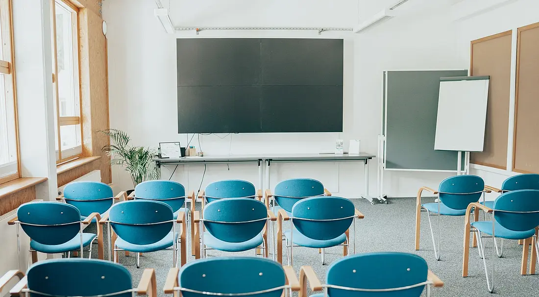 Blick in ein Tagungsraum: blaue Stühle stehen in vier Reihen in Richtung Videoleinwand.