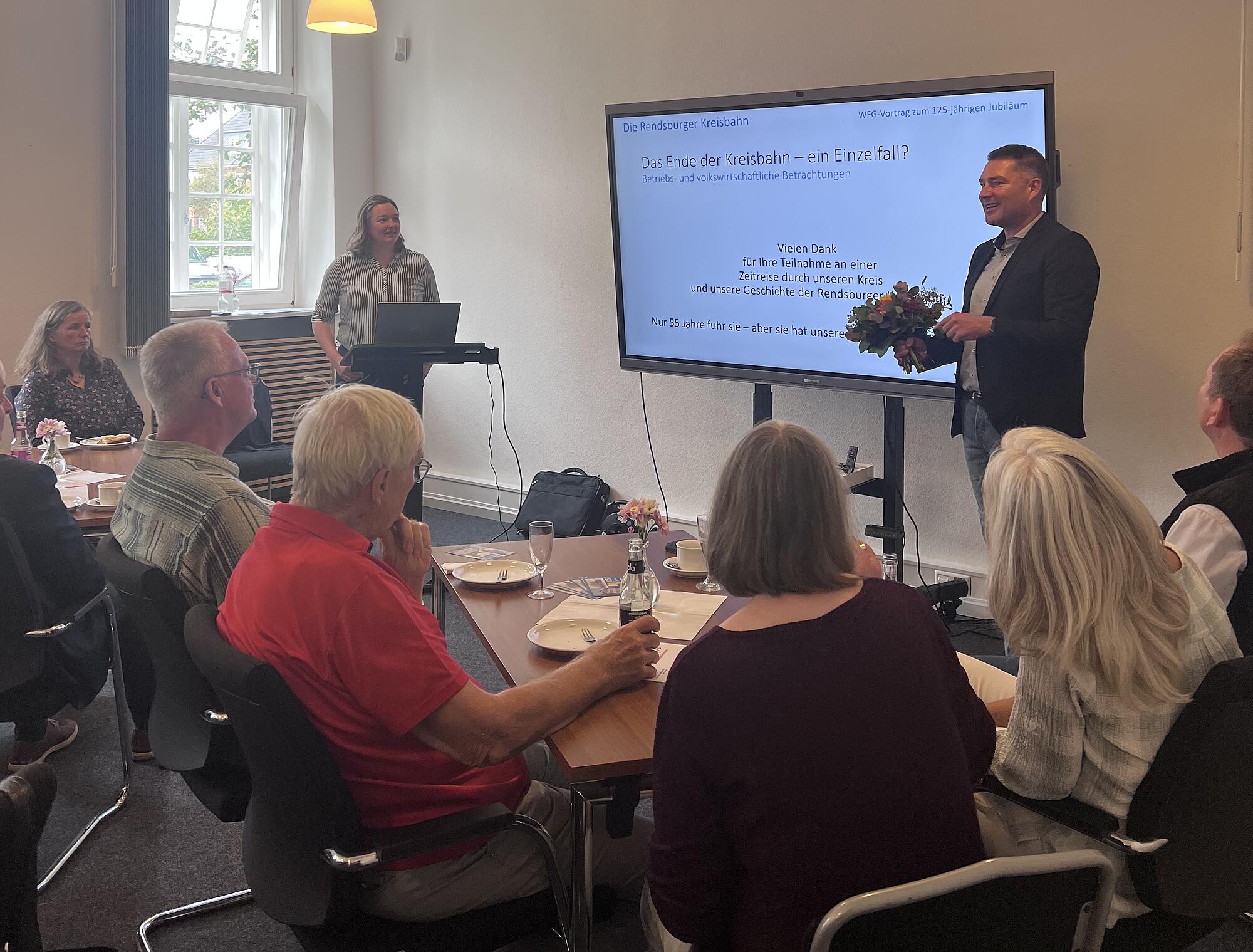 Ein Konferenzraum. Im Vordergrund sitzen Gäste und schauen auf einen Bildschirm, mit einer Präsentation. Davor ein Mann im Anzug mit Blumen in der Hand, links steht eine junge Frau am Rednerpult.