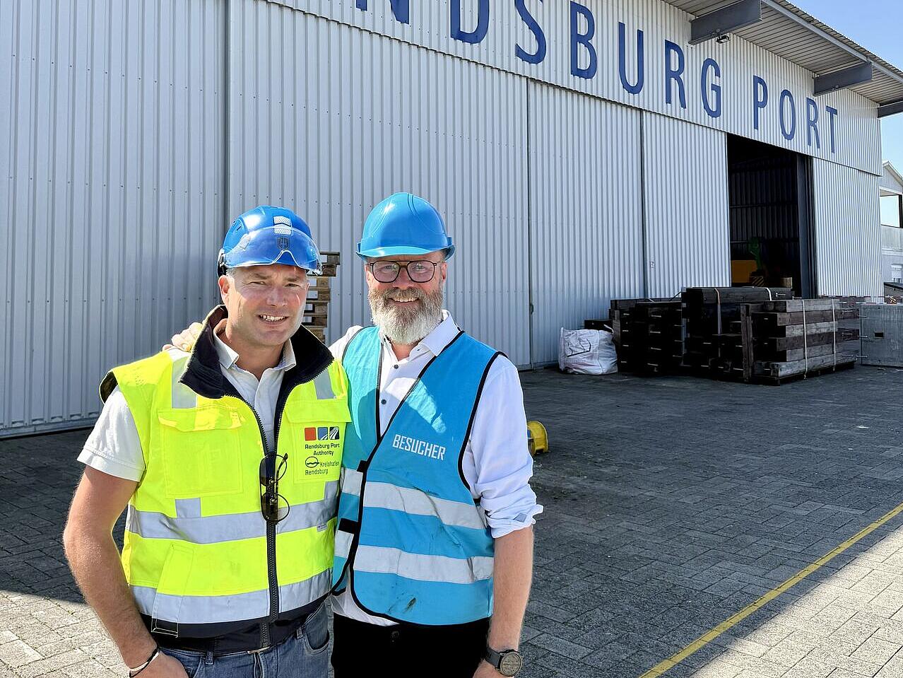 Wirtschaftsminister Claus Ruhe Madsen und Kai Lass, Geschäftsführer der Wirtschaftsförderung stehen mit Warnwetsten und Schutzhelmen vor der Werkshalle mit der Aufschrift "Rendsburg Port"