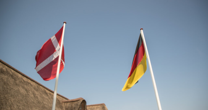 Eine dänische und eine deutsche Flagge an Fahnenstangen vor einem Reetdachhaus.