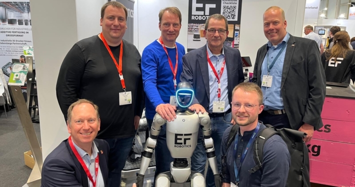 Sechs Männer auf einem Messestand. In der Mitte ein kleiner, weißer Roboter.