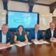 Vier Personen sitzen an einem Holztisch, vor ihnen Unterlagen, die sie offensichtlich unterzeichen wollen. Im Hintergrund ein großer Bildschirm auf dem das Logo von Clean Energy Valley zu sehen ist,