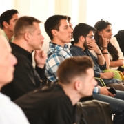 Mehrere Menschen sitzen in einer Veranstaltung in Reihen und hören offensichtlich einem Referenten, einer Referentin zu. Diesen sieht man aber nicht.