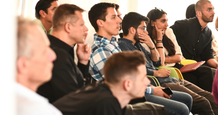 Mehrere Menschen sitzen in einer Veranstaltung in Reihen und hören offensichtlich einem Referenten, einer Referentin zu. Diesen sieht man aber nicht.