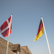 Man sieht das Dach eines Reet gedeckten Hauses, davor zwei Fahnenmasten in weiß. An dem einen hängt die dänische, an dem anderen die deutsche Flagge. Der Himmel ist wolkenlos blau.