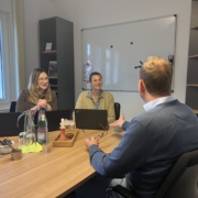 Zwei Frauen sitzen in einem Besprechungszimmer mit aufgeplappten Laptops vor sich. Sie reden mit einem Mann, der vor ihnen an einem Tisch sitzt. Dieser ist nur von hinten zu sehen.