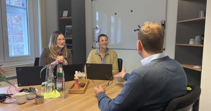 Zwei Frauen sitzen in einem Besprechungszimmer mit aufgeplappten Laptops vor sich. Sie reden mit einem Mann, der vor ihnen an einem Tisch sitzt. Dieser ist nur von hinten zu sehen.